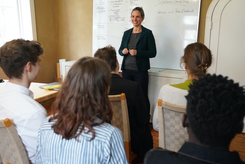 Pamela vor Whiteboard referierend mit jungen Menschen im Publikum